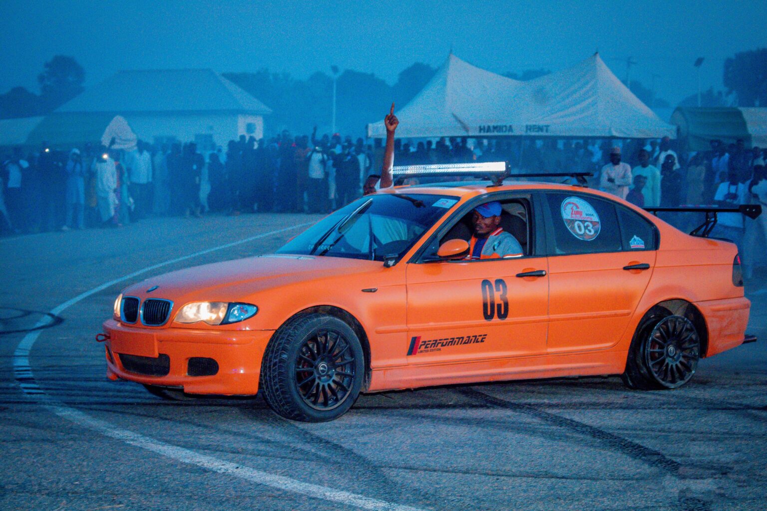 Bright orange car drifting during nighttime racing event with enthusiastic crowd.